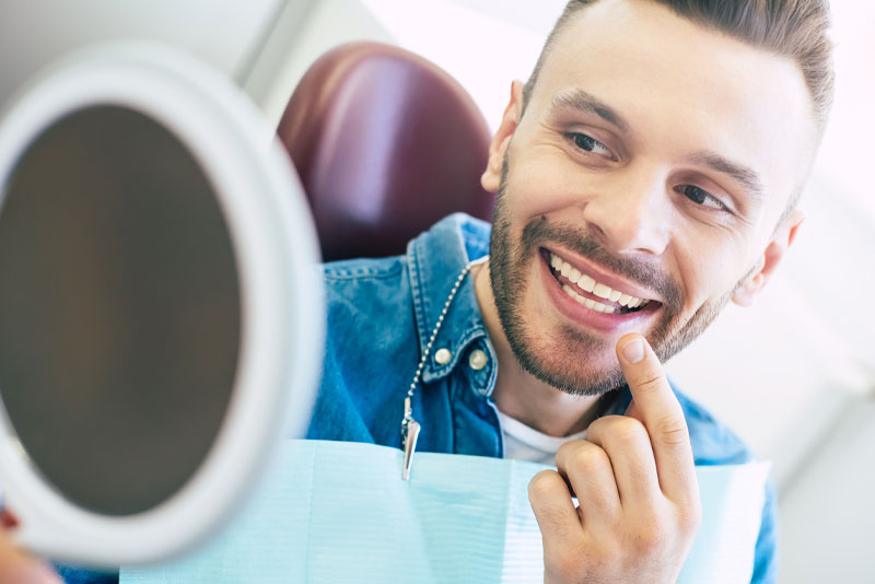 a dental patient smiling and pointing a finger at his cosmetically enhanced new smile, through a handheld mirror, after his smile makeover procedure.